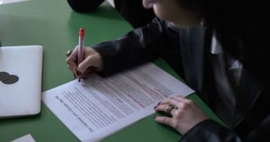 A person in a black jacket marks up a printed page with a red pen at a green desk, beside a closed laptop