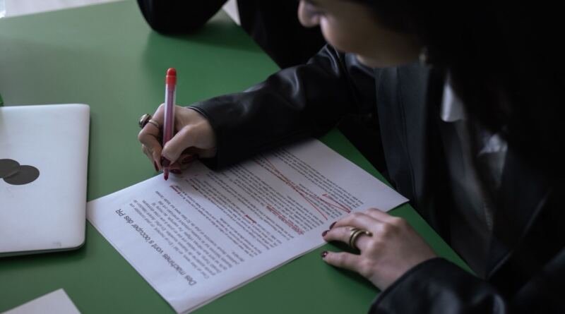 A person in a black jacket marks up a printed page with a red pen at a green desk, beside a closed laptop