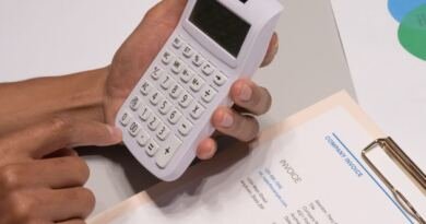 Two hands hold a small white calculator above a printed company invoice on a clipboard, with a gold pen resting beside it.