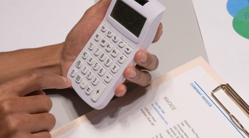Two hands hold a small white calculator above a printed company invoice on a clipboard, with a gold pen resting beside it.