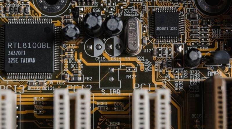 Close-up of a green computer motherboard, with a black RTL8100BL chip, silver capacitors and cream PCI slots on orange copper traces.