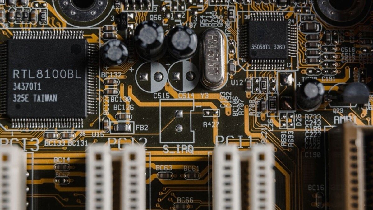 Close-up of a green computer motherboard, with a black RTL8100BL chip, silver capacitors and cream PCI slots on orange copper traces.