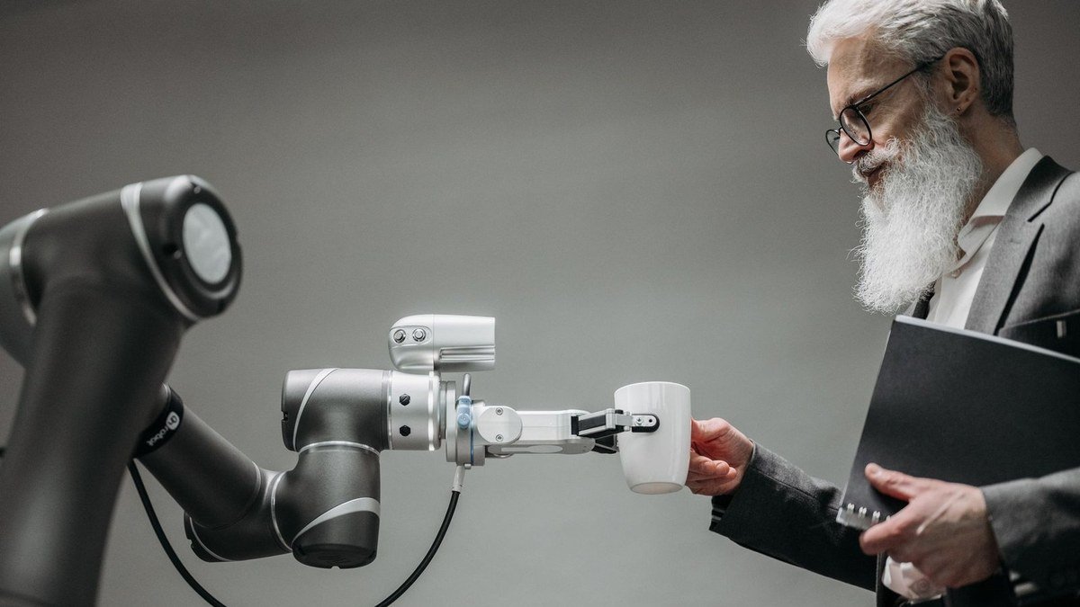 A grey-bearded man in a suit passes a white mug to the gripper of a robotic arm in a grey studio