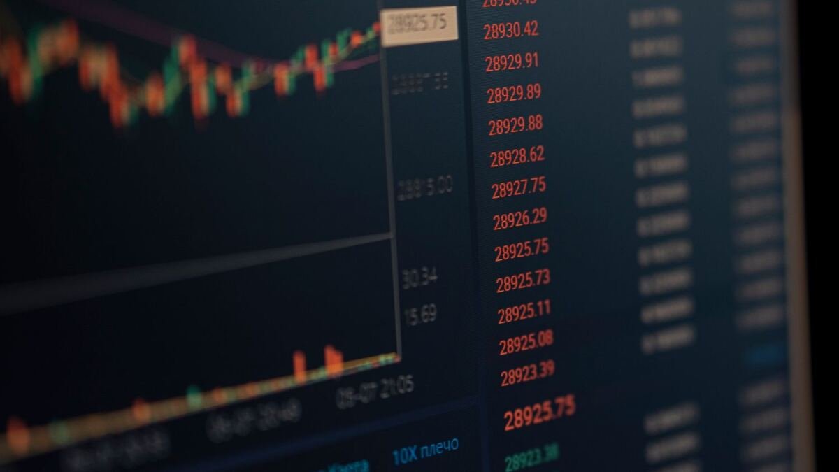 Close-up of a dark trading monitor showing a column of red price quotes beside a candlestick chart, photographed at an angle so the right edge blurs out of focus.