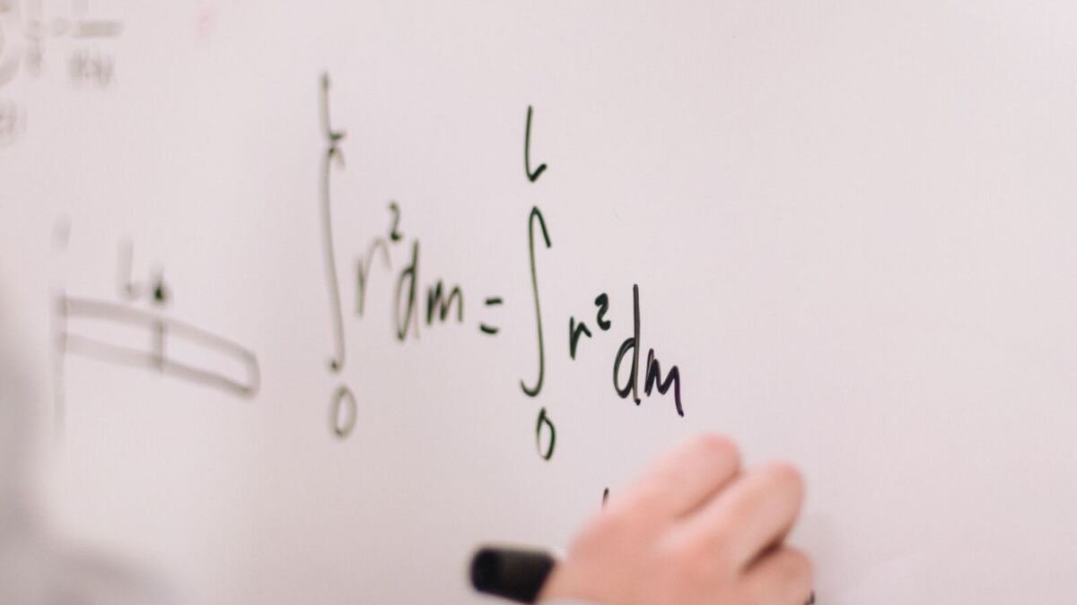 Close-up of a hand holding a black marker, writing integral equations in green and black ink on a white whiteboard.