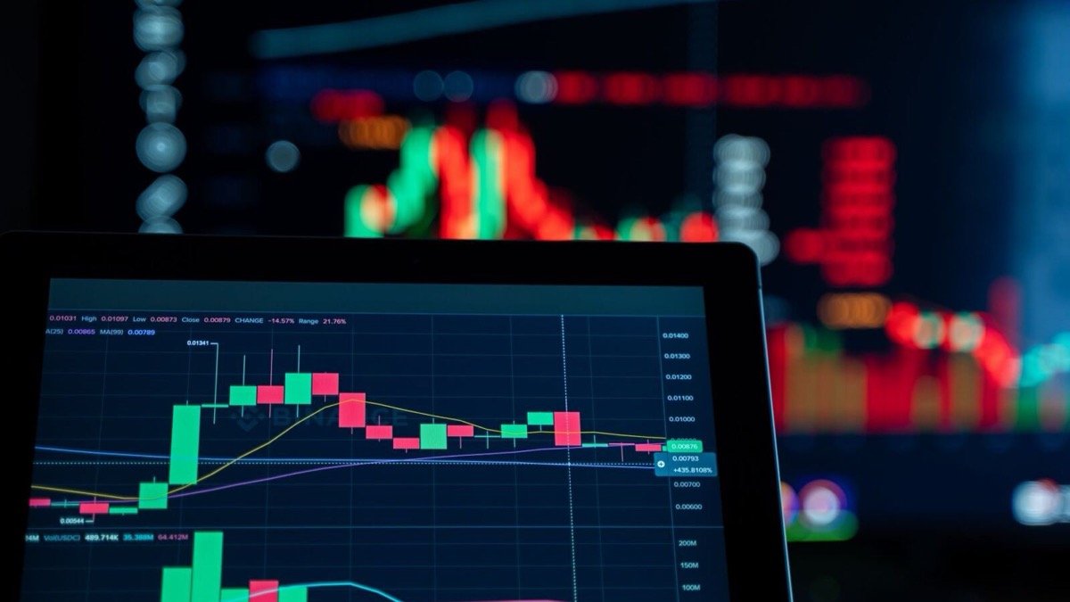 Laptop showing a candlestick trading chart, with blurred red and green market displays on a screen behind it