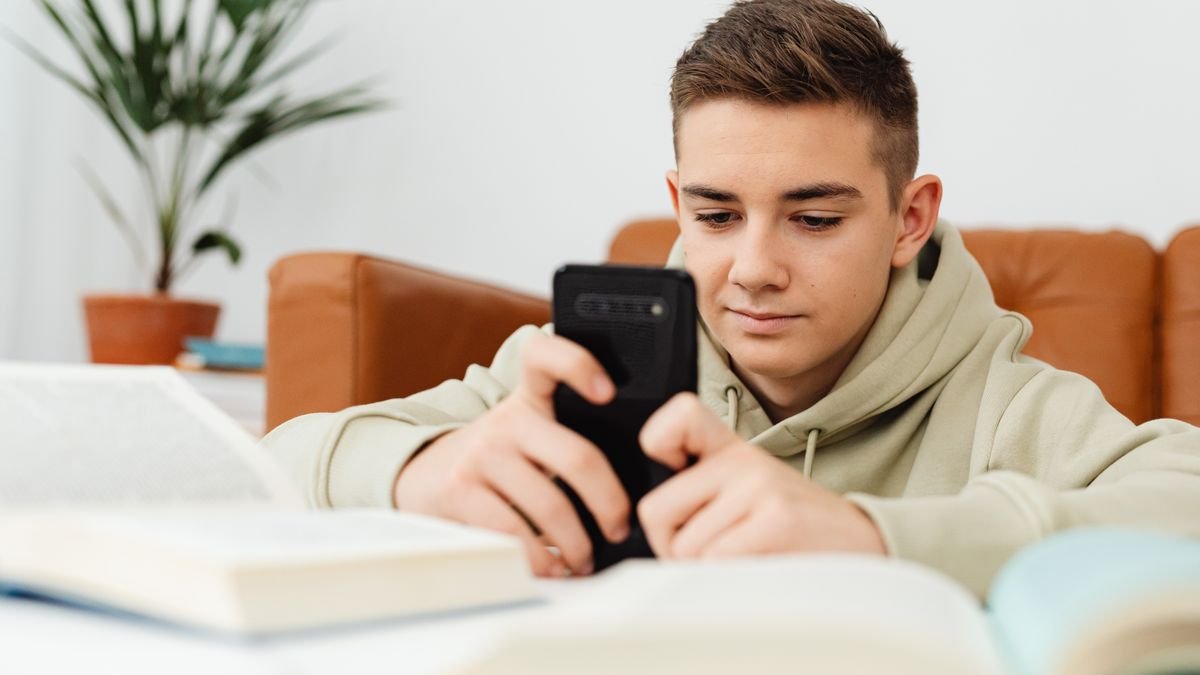 A teenager in a hoodie looks at a smartphone at a desk surrounded by books.