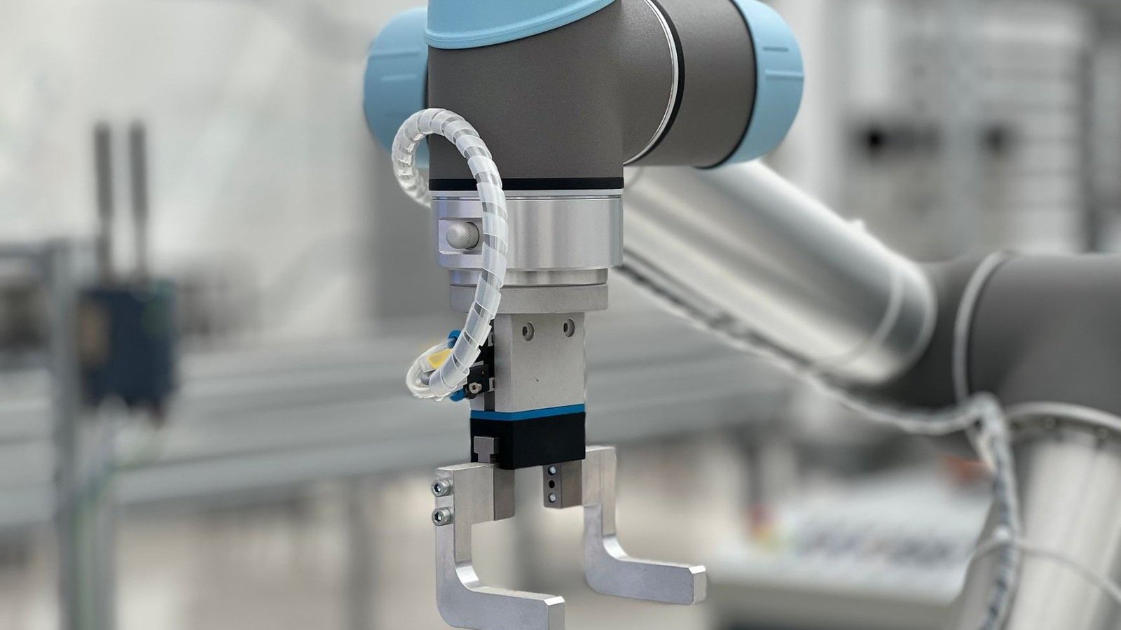 Close-up of a grey and light blue industrial robot arm with a metal two-finger gripper, in a blurred factory workspace
