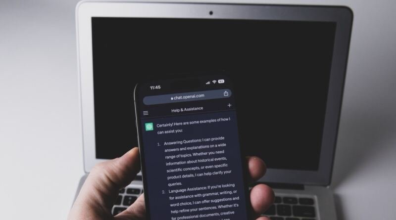 A hand holds a smartphone running one of the AI apps, a chatbot reply filling the screen, with an open laptop out of focus behind it