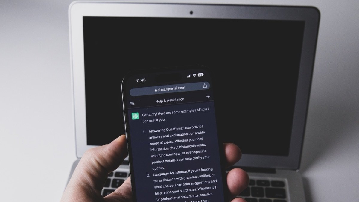 A hand holds a smartphone running one of the AI apps, a chatbot reply filling the screen, with an open laptop out of focus behind it
