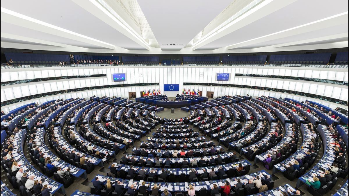 Rows of curved wooden desks and blue seats in the European Parliament hemicycle in Strasbourg during a plenary session, seat of the legislature that has AI defined in binding law.