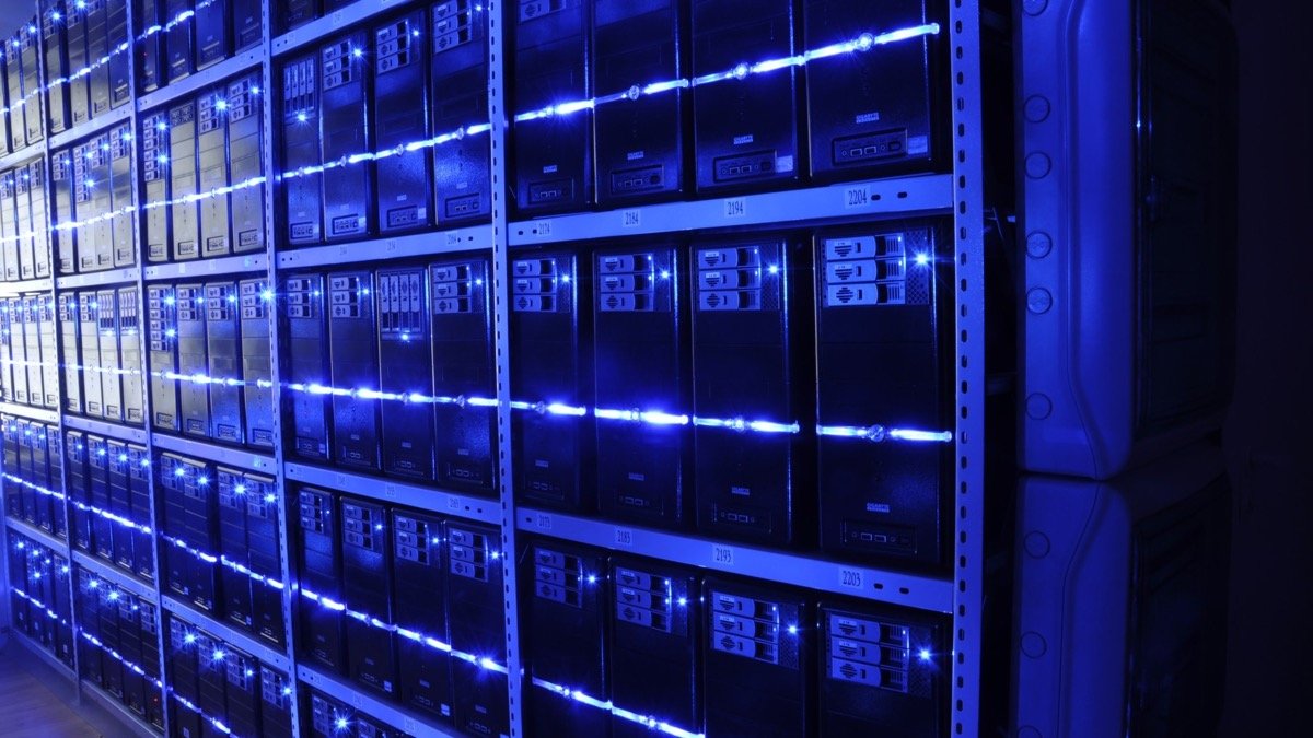 Rows of rack-mounted servers stacked on metal shelving in a darkened server room, each machine lit by a horizontal line of blue status LEDs