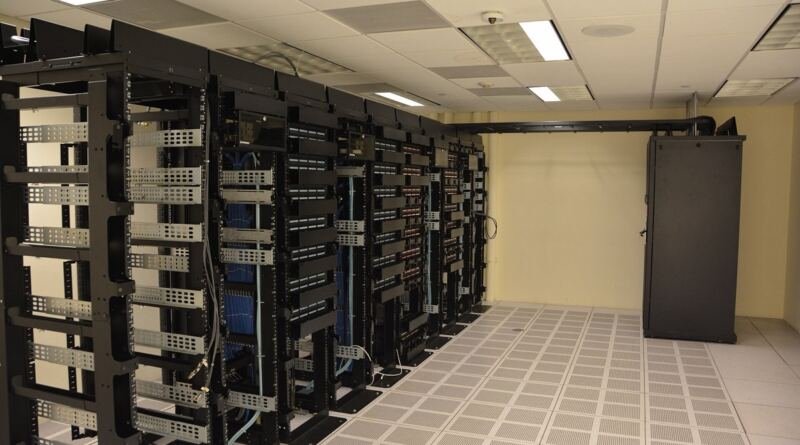 A row of tall black server racks with cable management arms and blue patch cables in a data centre, illustrating the hardware layer of the ai technology stack