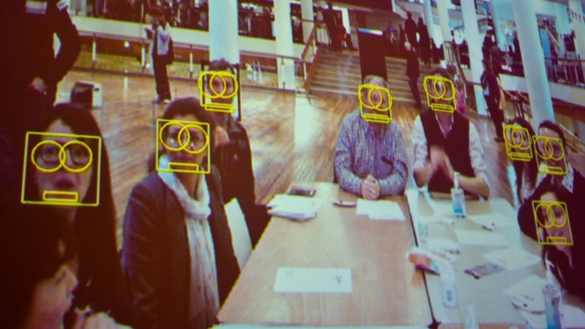 Projected camera feed showing yellow face-detection boxes over some but not all of the people seated at a long table, illustrating AI bias in facial analysis