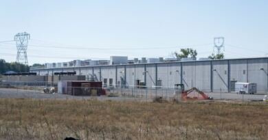 Long concrete data center building behind a chain link fence with power pylons overhead, illustrating the Amazon Data Center Commitment