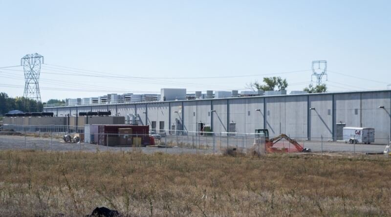 Long concrete data center building behind a chain link fence with power pylons overhead, illustrating the Amazon Data Center Commitment