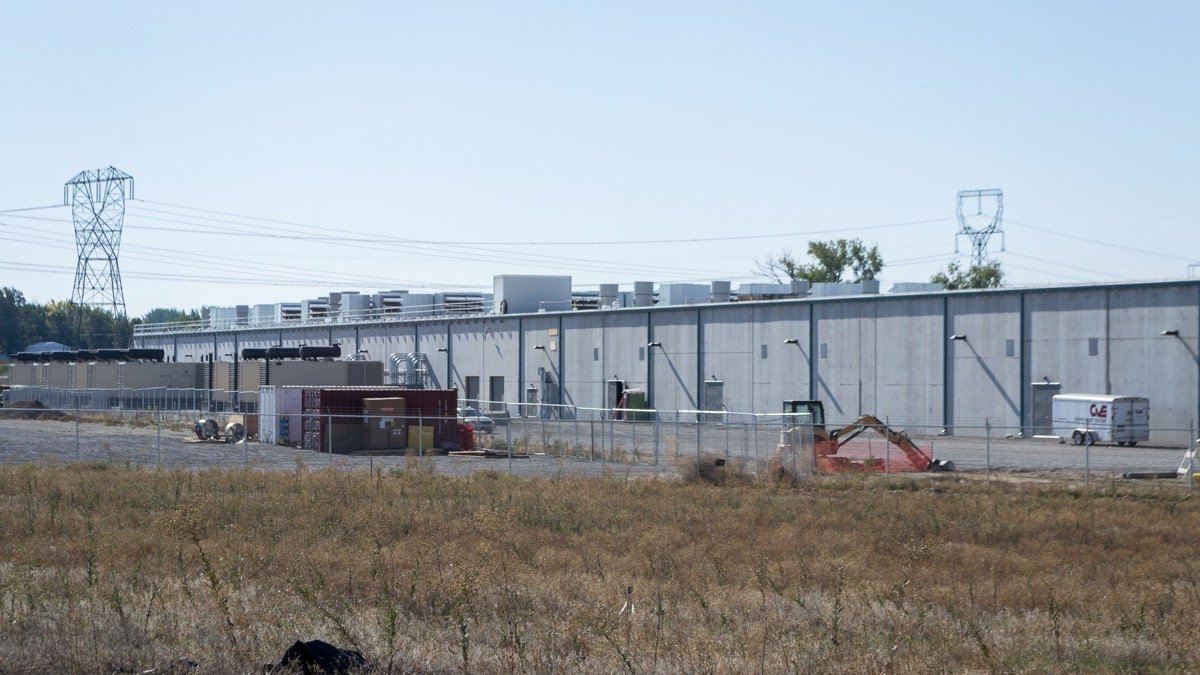 Long concrete data center building behind a chain link fence with power pylons overhead, illustrating the Amazon Data Center Commitment