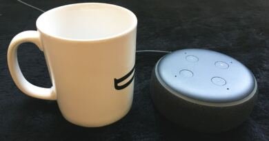 A white ceramic coffee mug stands beside a charcoal and grey Amazon Echo Dot smart speaker on a dark textured surface, a thin power cable trailing away behind them, illustrating AI in daily life at household scale