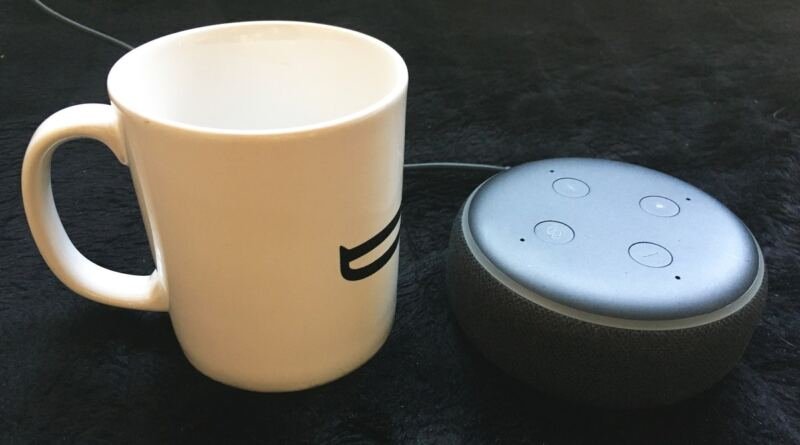 A white ceramic coffee mug stands beside a charcoal and grey Amazon Echo Dot smart speaker on a dark textured surface, a thin power cable trailing away behind them, illustrating AI in daily life at household scale